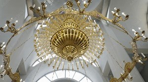 A magnificent golden chandelier adorns the white, curved ceiling of a church, its intricate design and lit candles embodying a blend of opulence and sanctity.