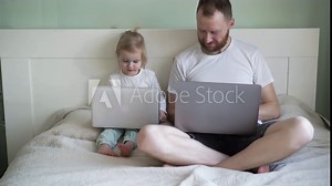 A father teaches his daughter to use a desktop laptop. Dad shows a small child how to perform actions on a computer. Sitting on the bed in a light interior in white tones.