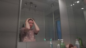 Anxious woman looking at her own reflection at bathroom mirror feeling regret and preoccupation. Female with emotional distress rubbing face and hair while looking at reflection in bathroom mirror.