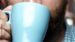 Bearded man is drinking coffee or tea. Male hand brings blue mug to her mouth and drinks coffee or tea from it