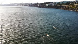 Aerial view of swimmers practicing with floats in the open ocean, swimming long distances. The wide shot captures the vast water and their determined effort to complete the challenging swim