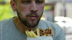 Traditional Greek street food - chicken gyros with sauce, vegetables and French fries wrapped in pita. Young guy eats a gyros in spring park on sunny day. Middle eastern street food cuisine