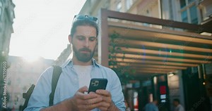 Attractive young man in good mood smiling, walking on city streets, holding smartphone in hands. Cheerful positive bearded male traveler using cellphone, scrolling, texting funny messages.