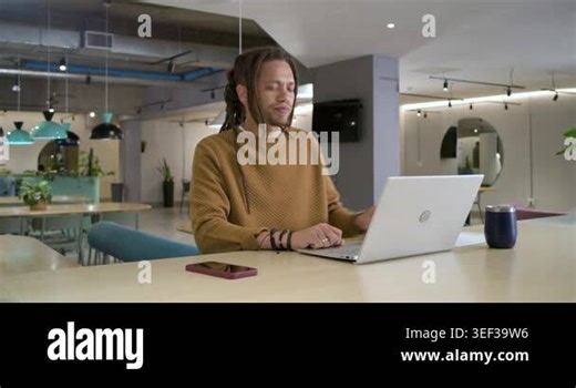 Man sipping blue mug, typing on laptop at shared table, checking pink phone after alert. Coworking, computer, monitor, plants, teal, pastel, modern Stock Video Footage - Alamy