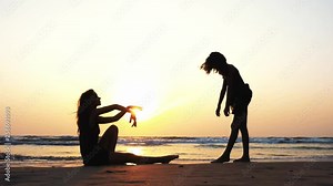Silhouette of a sporty woman trains a young girl to perform an acrobatic element of rhythmic gymnastics on the beach at beautiful sunset in slow motion. Side view.