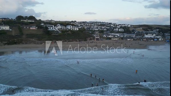 Aerial footage captures Polzeath Beach with gentle waves, distant village, and a serene twilight sky. Beachgoers enjoy the evening atmosphere, illuminated by soft sunset hues.
