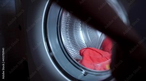 Person hands loading washing machine cylinder with dirty laundry, throwing transparent liquid yellow capsule inside and closing machine door. Housewife preparing dirty clothes for washing, routine