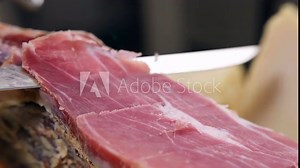 Professional chef slicing cured prosciutto leg with sharp knife, extra quality Italian or Spanish jamon leg for typical mediterranean antipasto, dry-cured prosciutto slices close-up