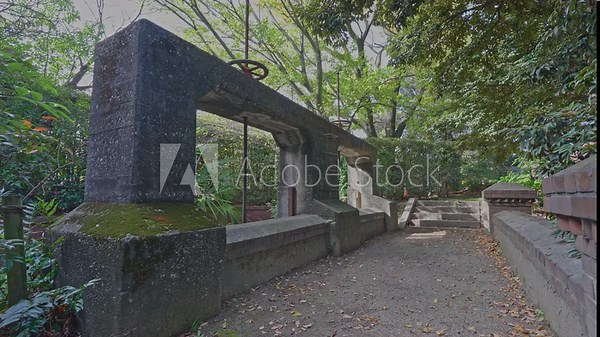 Historic concrete irrigation weir gate (Jinzaemon Weir) with a visible valve wheel, surrounded by overgrown park greenery.