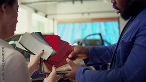 Salesperson in auto showroom discussing with man, offering color swatch samples choice. Agent providing client with color wheel selection, picking hue for car paint, camera B close up