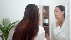 A woman in a bathrobe, at home, explores a beauty skincare concept, gazing into the bathroom mirror with care and intention.