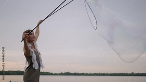 A young female artist shows a soap bubble show blowing up huge soap bubbles on the shore of a lake at sunset. Show a beautiful show of soap bubbles in slow motion