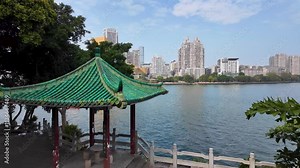 A traditional Chinese pavilion with an ornate roof structure stands on the riverbank of the scenic Liujiang River, surrounded by modern high-rise buildings of the urban skyline. City of Liuzhou, China