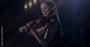 Caucasian female violinist in elegant look performing music on stage in darkness and spotlight like virtuoso. Beautiful woman violinist playing on violin at classical concert.