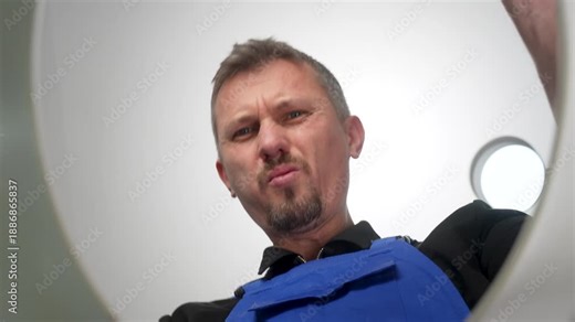 Plumber in a blue worker uniform pinches his nose while looking down, reacting to a bad smell. View from inside a toilet, representing plumbing issues, sewage problems, or cleaning services