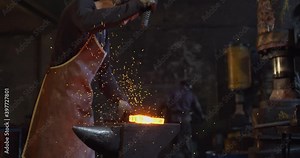 A blacksmith in a leather apron with a face in soot stands by the anvil and strikes the red-hot metal with a powerful hammer, causing a huge column of fiery sparks. High quality 4k footage