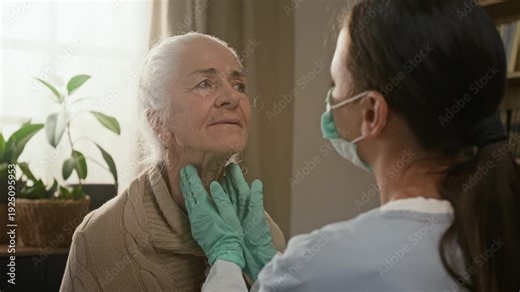 Over the shoulder rack focus shot of brunette young nurse wearing rubber gloves palpating lymph nodes of senior ladys neck during home medical examination