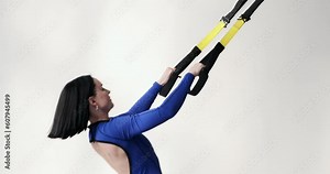 Confident Caucasian woman wearing a blue athletic jumpsuit and sports shoes doing pull-ups with resistance bands. She pulls herself up using the loops.