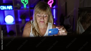 Blonde, middle age woman streamer in gaming room feverishly buying gear with credit card on computer, immersed in the virtual stream of her thrilling gaming night.
