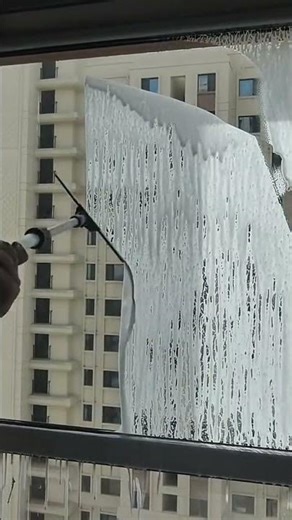Cleaning building windows with soapy foam using a long telescopic pole from inside an apartment unit