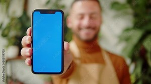 Forist holding phone with chroma key. Greenhouse worker portrait. Arabian man botanist smiling, small business owner. Gardener working in flower shop, plant store.