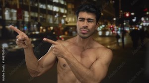 Hispanic shirtless man showing bare chest pointing fingers amid neon lit urban street at night; indecision.