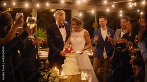 Beautiful Bride and Groom Celebrate Wedding at an Evening Reception Party with Multiethnic Friends. Married Couple Standing at a Dinner Table, Kiss and Cut Wedding Cake.