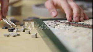 Male hands create mosaics pattern using tweezers and small pieces of granite stone. Master making handicraft in creative workshop. Interesting hobby, art, artwork. Close up. Slow motion ready 59.94fps