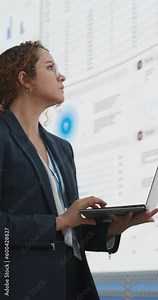 Vertical Screen: Hispanic Businesswoman Holding Laptop Computer And Analyzing Data On Big Digital Screen With Graphs and Charts in Monitoring Office. Female IT Entrepreneur Running Software Company.