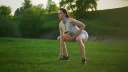 A woman jumps up and squats in the park at sunset. Training a young...