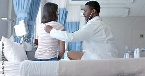 Diverse male doctor using stethoscope on back of female patient in hospital ward, slow motion
