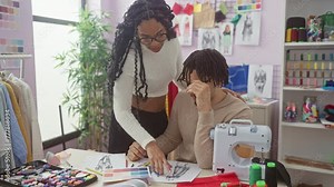 A woman and a man collaborate in a tailoring atelier among sewing machine, fabrics, sketches, and thread