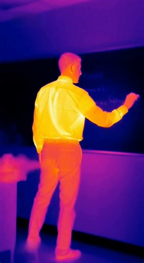Thermal Imaging Turns Ordinary Lecture Into Unplanned Comedy Classroom setting, daytime — What should have been a straightforward classroom moment ends up becoming unexpectedly memorable when thermal imaging reveals more than anyone intended. The clip shows a professor standing near a large presentation screen, explaining a point while gesturing with both hands. Through the thermal filter, the room glows in layered colors—cool blues along the walls, warmer tones outlining the professor’s body an