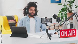 Young enthusiastic Arabian man blogger shoots video on mobile phone camera and applauds announcing results competition on page on social networks sits at table with microphone in home office. On air