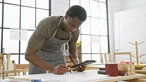 Focused man using tablet while drafting plans in a bright workshop, wearing an apron and surrounded by woodworking tools.