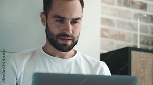 A smiling young man is using his laptop computer sitting indoors