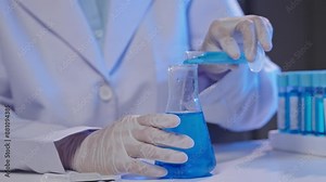 Scientist in lab coat pours blue liquid from test tube into flask while conducting experiment. Gloves and scientific glassware visible