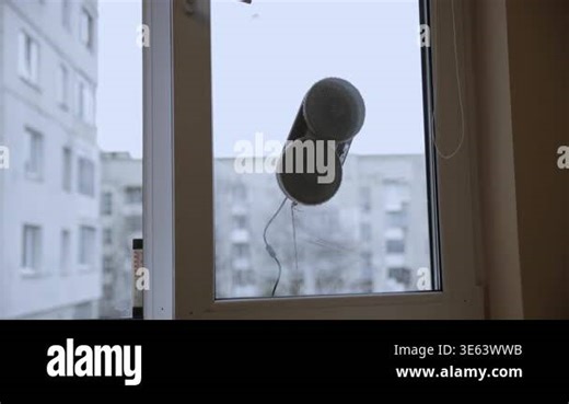 An automatic window cleaning robot with grey microfiber pads working on a glass pane. Background shows blurred city residential buildings under a grey sky. Modern smart home technology for high-rise apartment maintenance and effortless chores Stock Video Footage - Alamy
