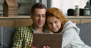 Happy young couple has video chat with their friends, family members, holding tablet computer, smiling, looking at webcamera, sitting on grey sofa at home. 4K, shot on RED camera.