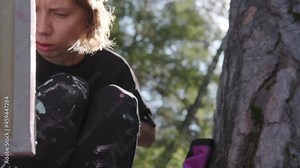 Modern stylish girl artist paints a picture in the forest, leaning a canvas on a tree. Focused on the process, frowns and creates. Young creative free woman artist paints in nature on canvas.