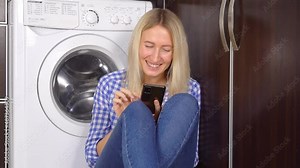 a happy modern woman using a home cleaning app on a smartphone in a modern home. She communicates via messenger, sitting by the washing machine on the kitchen floor. smart home,