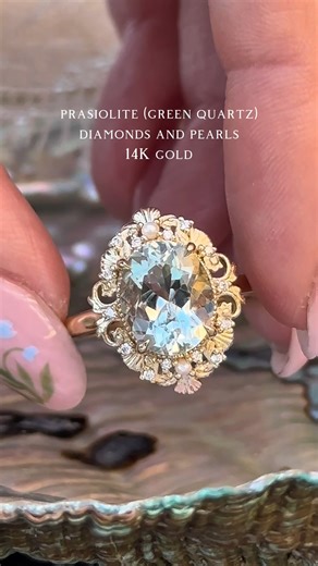 a ring inspired by mermaids and the birth of venus, introducing the “Merrow” ring 🌊˖°𓇼⋆🧜‍♀️🐚 𓈒𓏸 shown in yellow gold with an oval prasiolite (green quartz) center stone, pearl and diamond accents, and delicate shell filigree #mermaidday #mermaidaesthetic #uniqueengagementrings #sirencore #jewelrydesigner | Staghead Designs