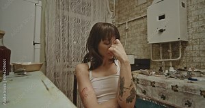 Wasted woman sitting in kitchen. Tattooed female junkie touching head and embracing knees while sitting on chair in grungy kitchen at home