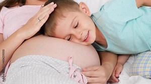 Cute little boy in pajamas listening to unborn baby in pregnant mother belly lying on bed. Concept of loving children and family happiness expecting baby