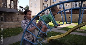 A two and a half year old boy is climbing a ladder in a playground. A babysitter is helping the baby climb up the ladder on the children's slide. Little feet climbing in shorts and sandals