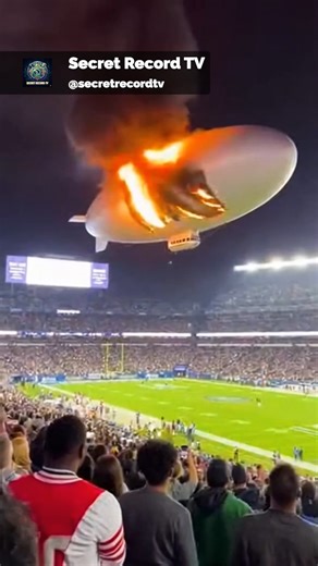 Fans cheering at a packed stadium are thrown into panic as a blimp soaring above suddenly catches fire. Flames and smoke trail across the sky, creating a terrifying spectacle as spectators duck and flee in fear. Emergency crews spring into action, directing the crowd to safety and working to control the blazing aircraft before it can crash onto the field. Cameras capture the dramatic scene from multiple angles, showing the blimp’s fiery descent and the frantic efforts of first responders. The in