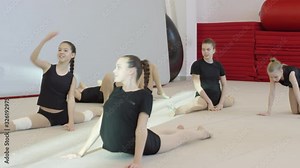 Tracking shot of cheerful teen and preteen girls in black t-shirts and shorts stretching on floor before cheerleading practice. They are doing gymnastic stretches for their legs, backs and hips
