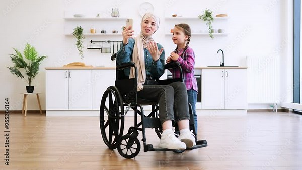 Bright, airy setting highlights family bonding, technology use, and inclusion. Muslim mother in wheelchair and daughter have video call, take selfie using phone in modern kitchen.