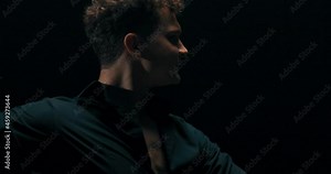 A professional experienced men is dancing on a dark stage lit by a lamp, practicing new moves, poses, the man has black shirt unbuttoned, he is a choreographer, showing steps to beginner dancers