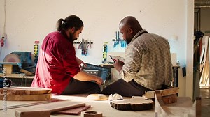 BIPOC artisan and apprentice comparing wood art piece schematic with CAD software 3D render on laptop. Woodworking experts in studio crosscheck drawing on clipboard with notebook blueprint, camera B